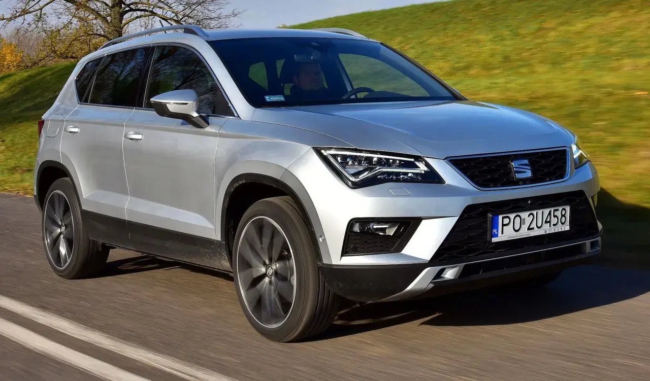 Srebrny Seat Ateca w ruchu. Kierowca za kierownicą, zielone wzgórza w tle. Wiele osób szuka seat ateca opinie przed zakupem.