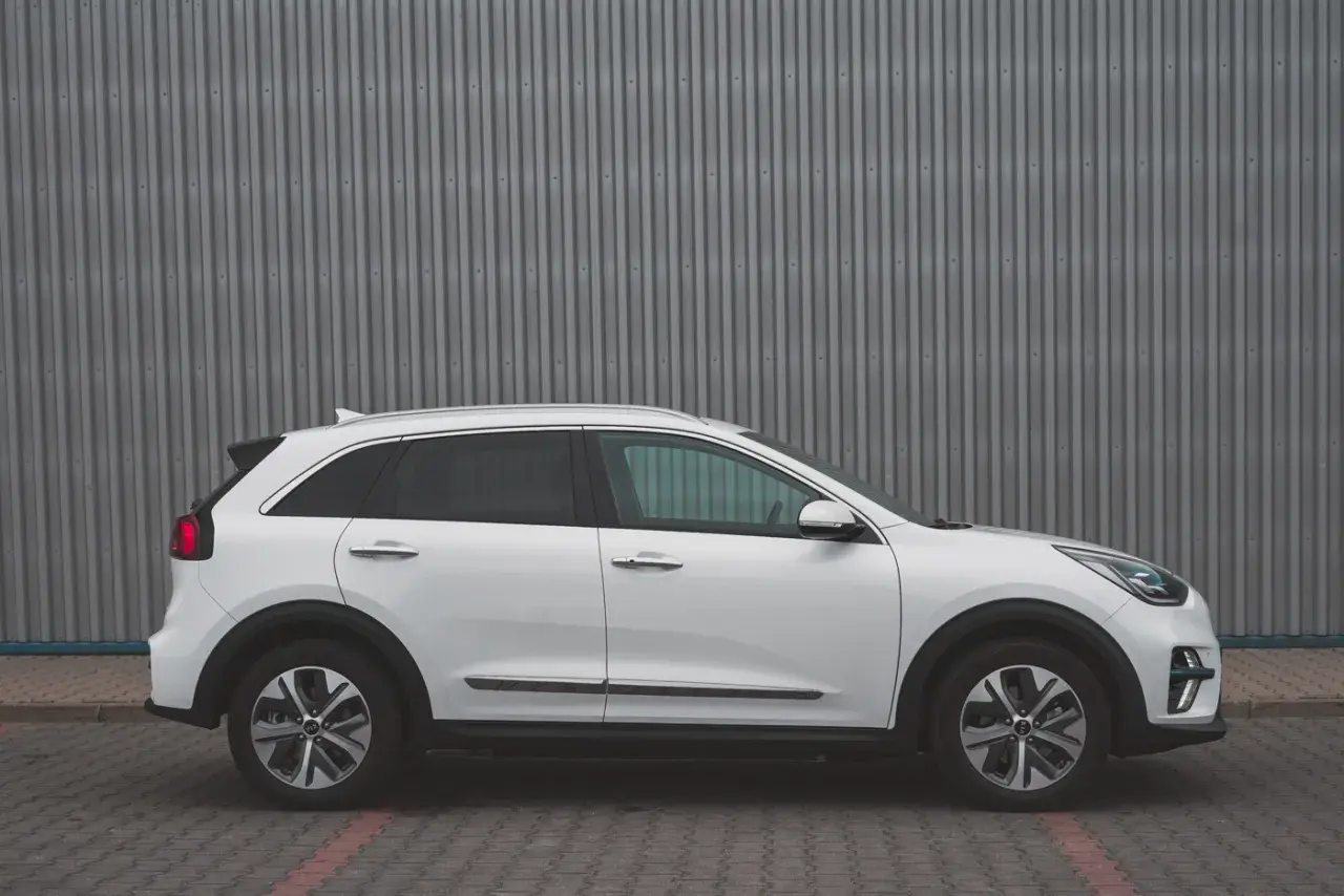 Biały Kia Niro EV stoi na tle szarej, falistej ściany. Elegancki design i nowoczesne linie podkreślają jego ekologiczny charakter.
