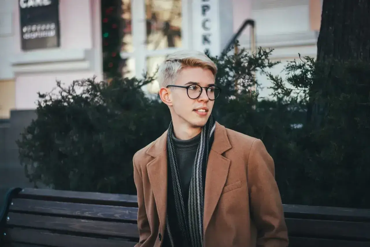 Młody mężczyzna w okularach i płaszczu prezentuje sportową elegancję, siedząc na ławce wśród zieleni.