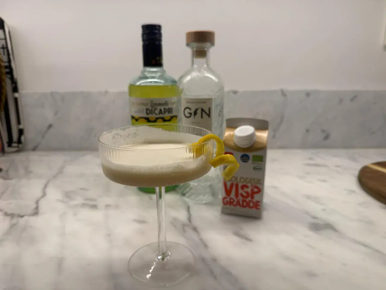 En krämig limoncello drink med citronskal, bredvid flaskor med limoncello, gin och vispgrädde.