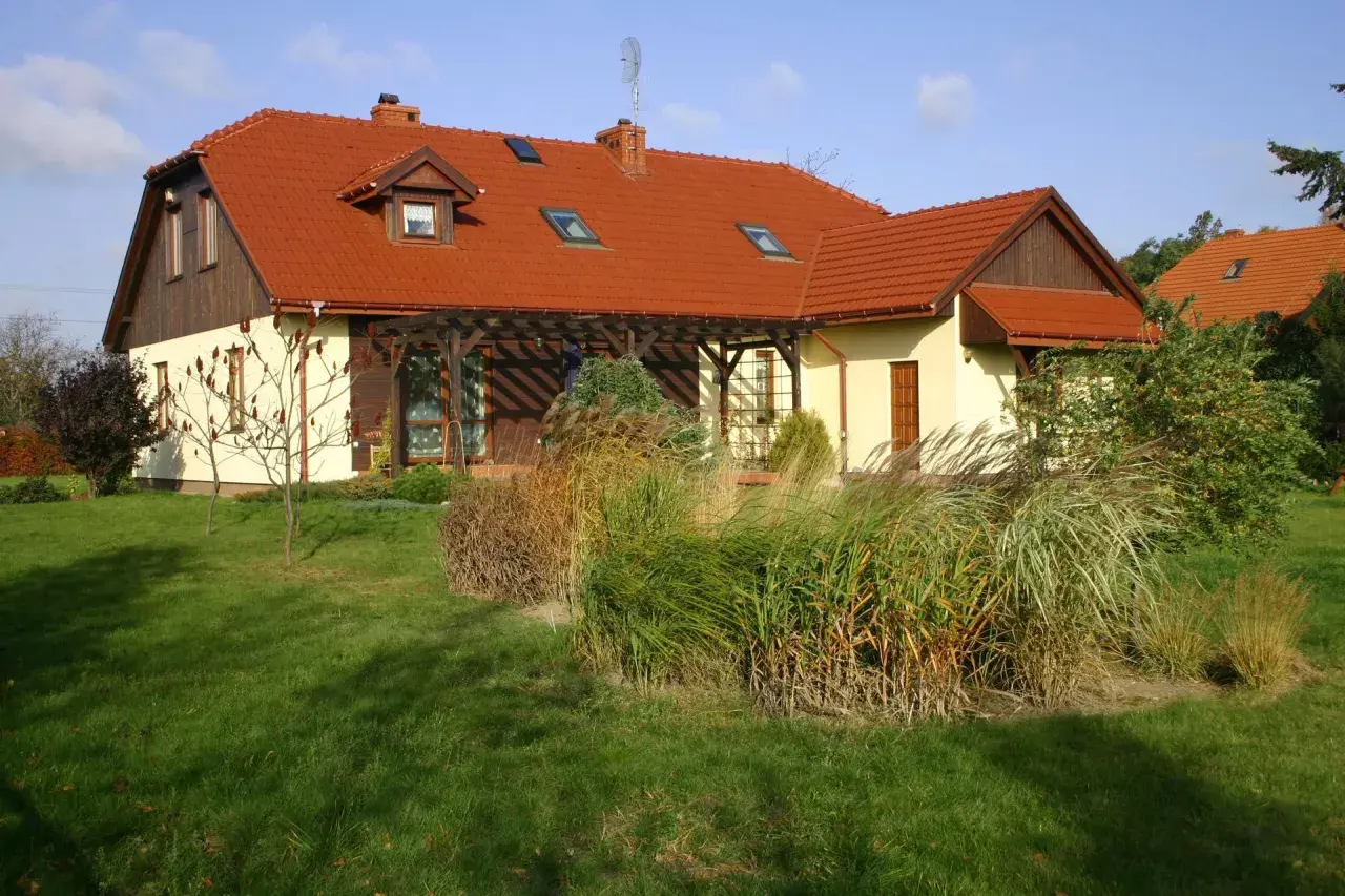 Dom z czerwonym dachem naczółkowym, z werandą i ogrodem pełnym traw ozdobnych.