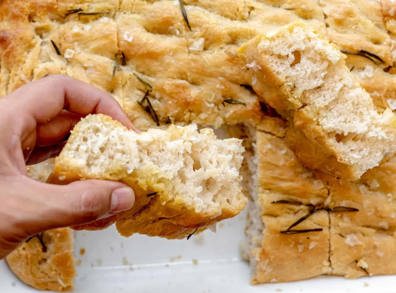 Mano rompe un trozo de focaccia original, dorada y con romero.
