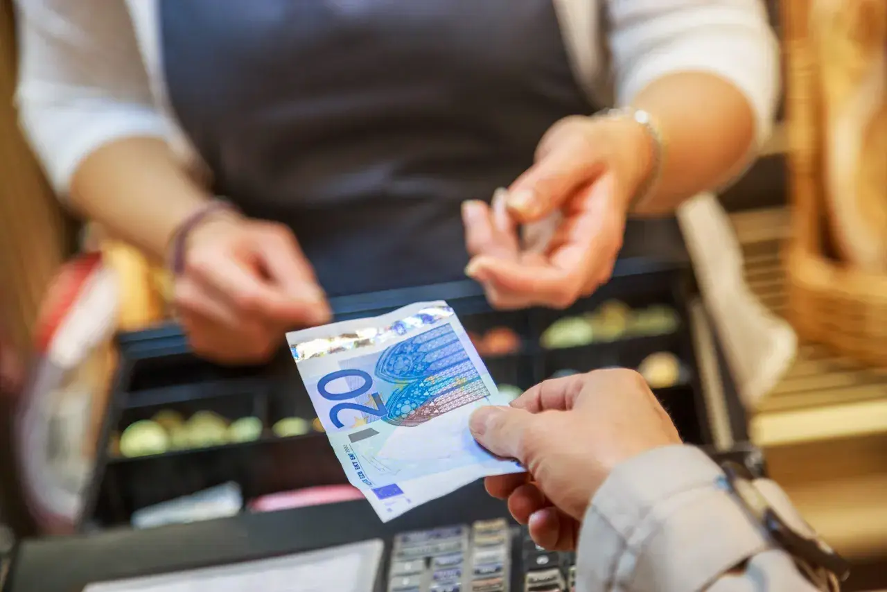 Une main tend un billet de 20 euros pour une saisie avoir espèces. Une caisse enregistreuse est visible en arrière-plan.