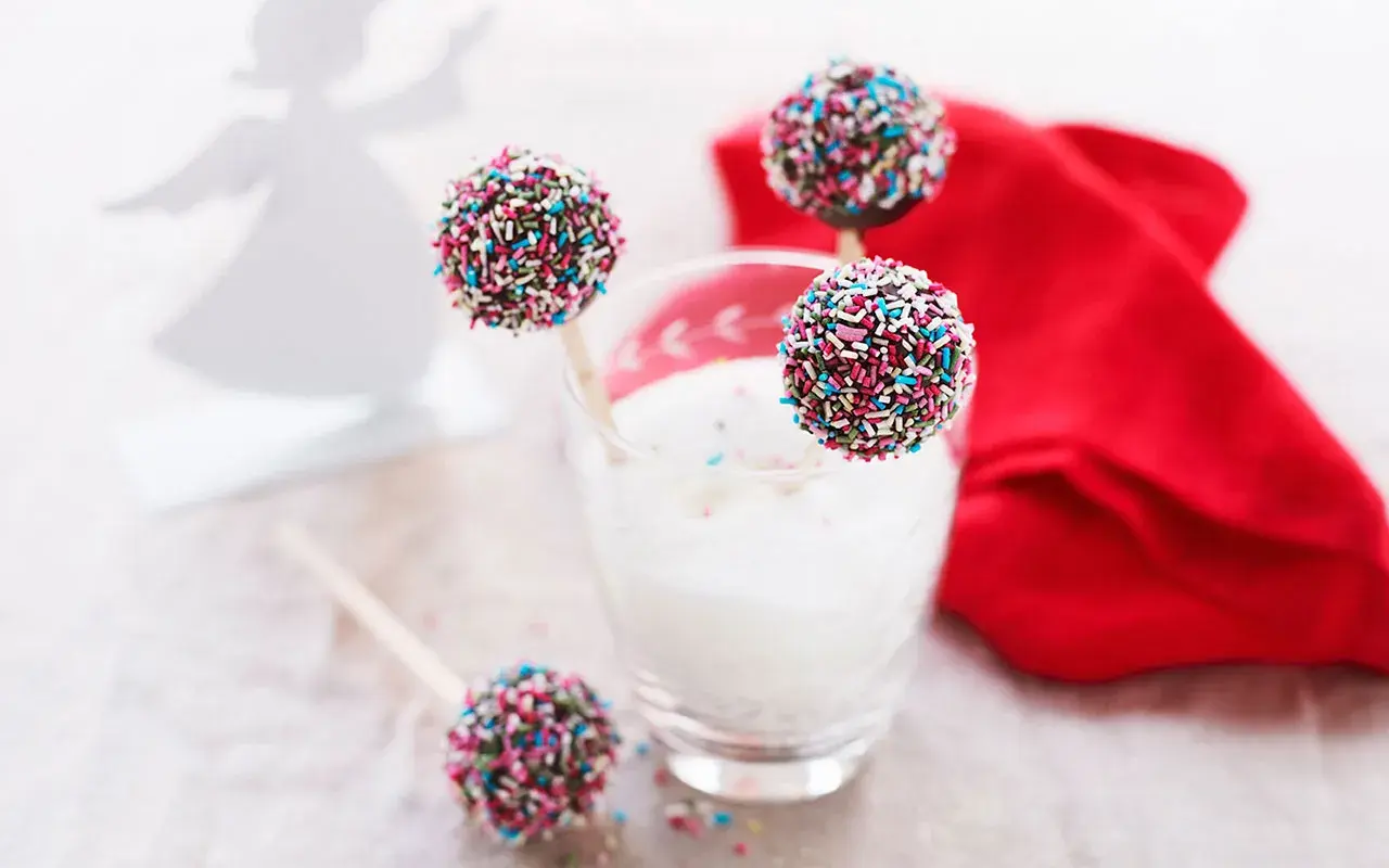 Färgglada cake pops med strössel står i ett glas med mjölk. En röd duk och en ängel i bakgrunden skapar en festlig stämning.