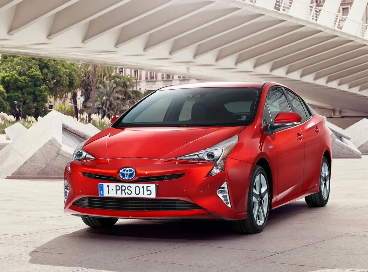 Czerwony Prius 4 z nowoczesnym designem, zaparkowany pod betonową konstrukcją z palmami w tle.