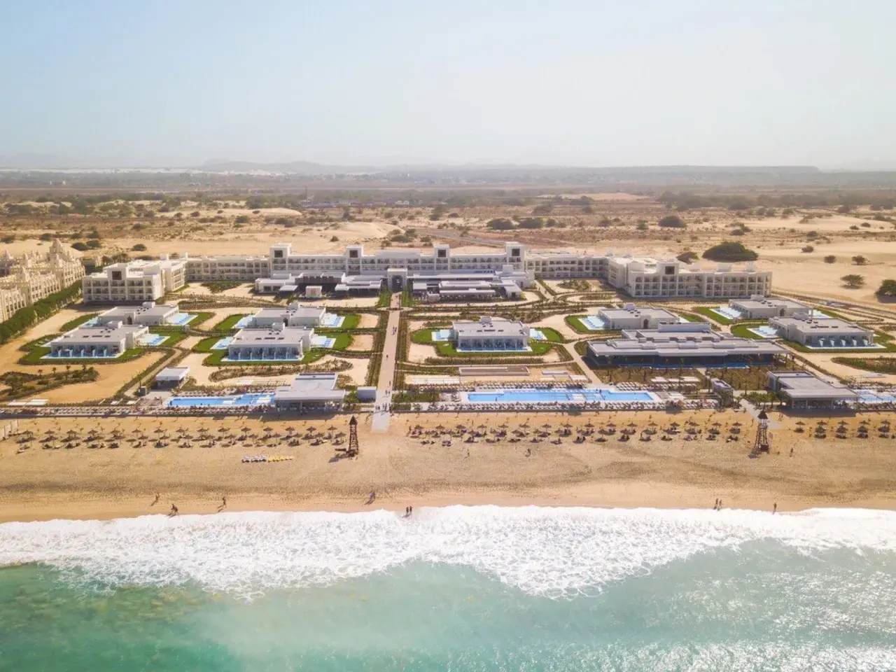 Luftaufnahme des Riu Palace Boavista Hotels mit mehreren Gebäuden, Pools und einem Strand mit Sonnenschirmen.