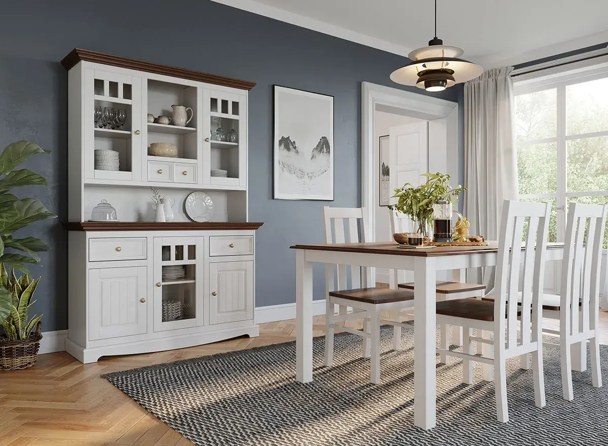 Biały kredens z brązowym blatem i detalami, pełen naczyń i kieliszków, stoi obok stołu z krzesłami w jadalni.