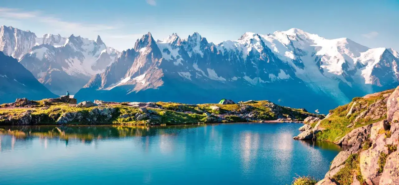 A serene lake reflects snow-capped peaks during a Mont Blanc trek. A tent sits on a grassy knoll, hinting at an overnight adventure.