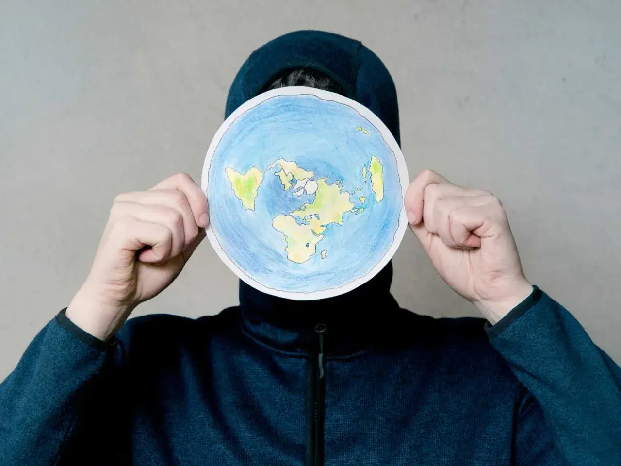 Osoba w kapturze trzyma okrągły rysunek płaskiej Ziemi. To nie jest globe po polsku, ale ciekawa wizja świata.