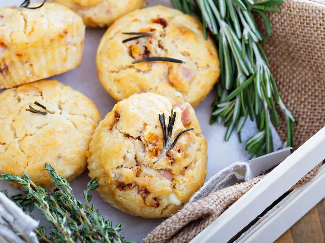 Vier herzhafte Muffins mit Rosmarin und Speck, garniert mit frischen Kräutern, liegen in einer Holzkiste.
