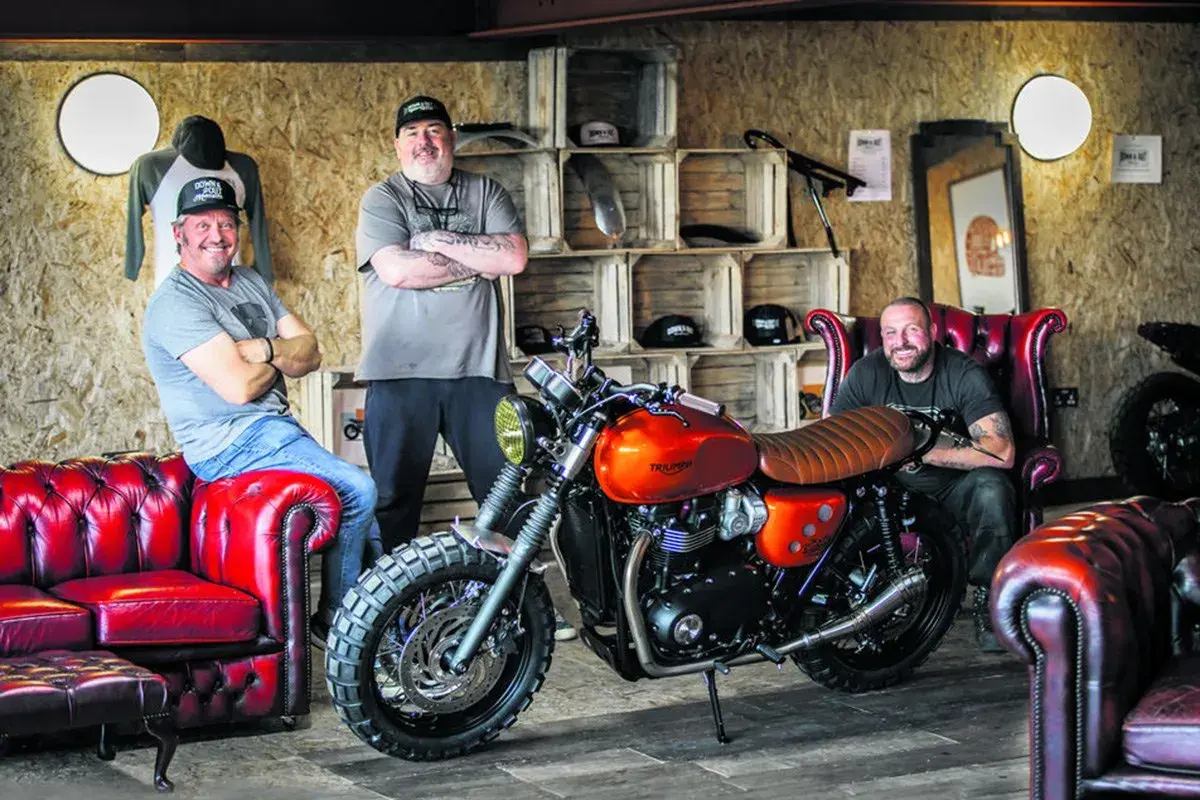 Three men pose with a custom orange Triumph motorcycle, showcasing the work of best custom bike builders.