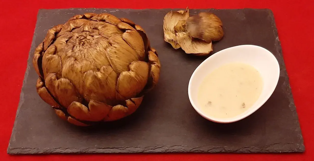 Un artichaut entier, cuit à la cocotte minute, servi avec sa sauce et quelques feuilles détachées sur une ardoise.