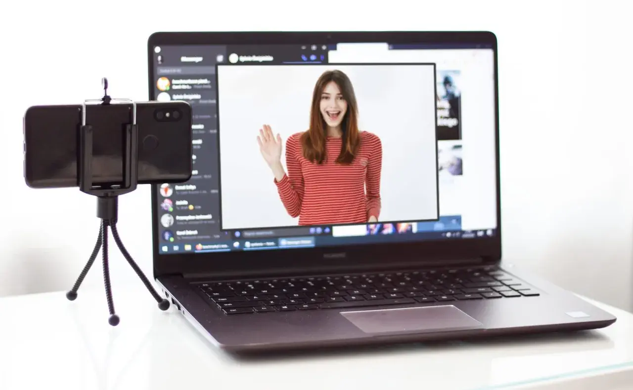 Kamera z telefonu na PC: młoda kobieta w bluzce w paski wita się na ekranie laptopa, obok stoi telefon na statywie.