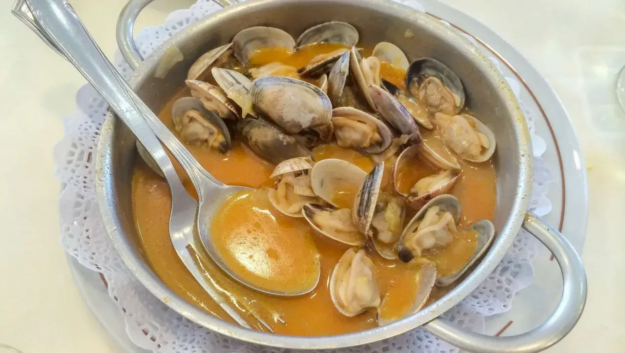 Almejas en una cazuela metálica, bañadas en una sabrosa salsa marinera. Una cuchara plateada reposa sobre el plato, lista para disfrutar.