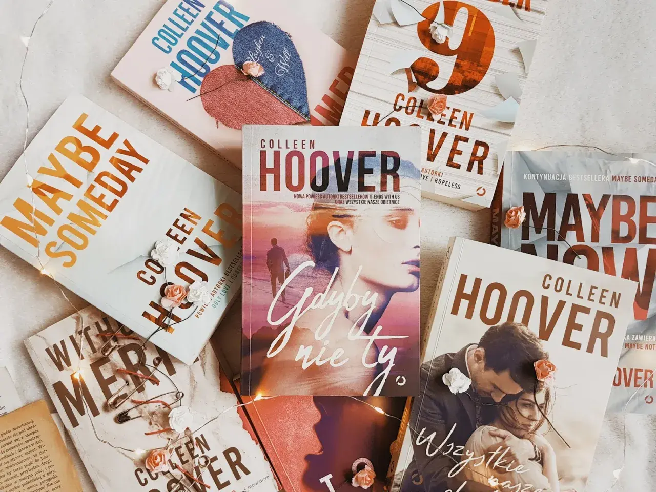 Książki Colleen Hoover ułożone na jasnym tle, ozdobione światełkami i sztucznymi kwiatami. Wśród nich "Gdyby nie ty", "Maybe Someday" i "Wszystkie nasze obietnice".
