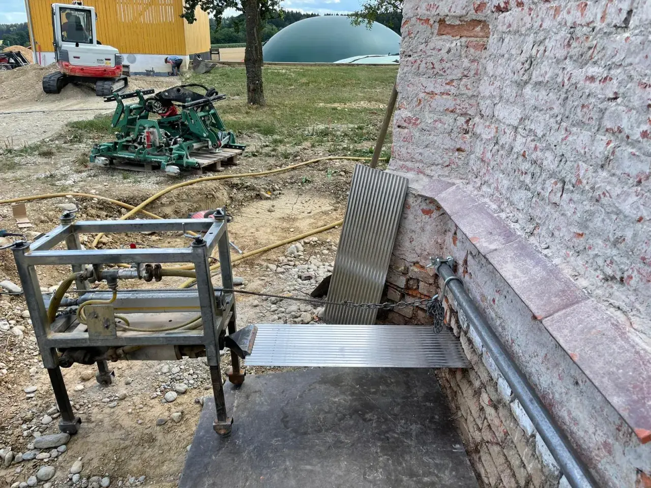 Baustelle mit Bagger, Maschine und Metallkonstruktion. Eine horizontale Sperrschicht aus Metallblech schützt die Ziegelwand.