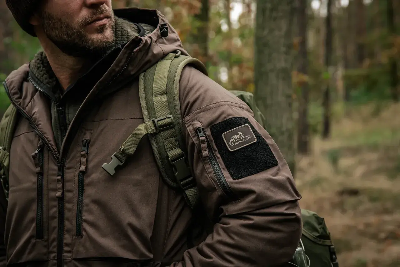 Mężczyzna w brązowej kurtce z naszywką Helikon-Tex, symbol polskie marki sportowe, w lesie.