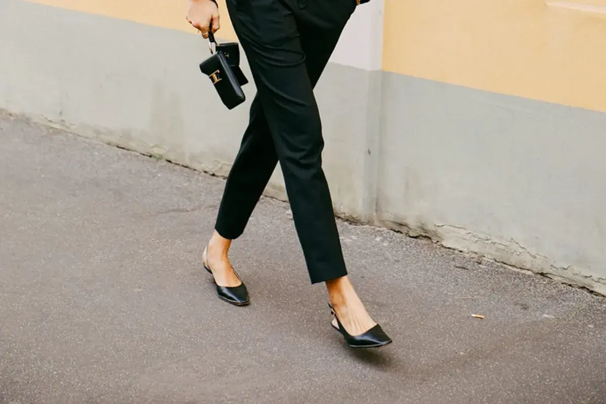 Une femme élégante marche dans la rue, portant un pantalon noir et des chaussures ballerines noires. Elle tient un petit sac noir.
