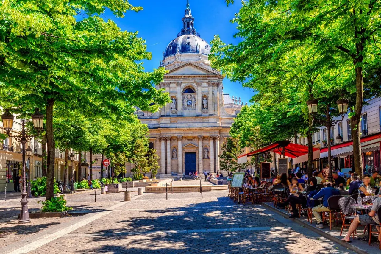 En solig paris april med gröna träd som ramar in en vacker byggnad. Folk sitter vid cafébord i skuggan.