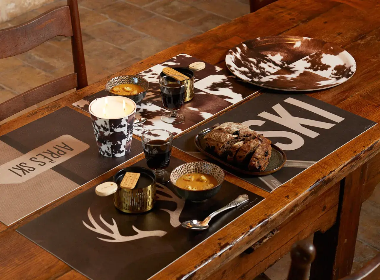 Ambiance chaleureuse pour une deco table raclette : bougie, bols de soupe, pain et verres de vin sur des sets "après ski" et motif vache.