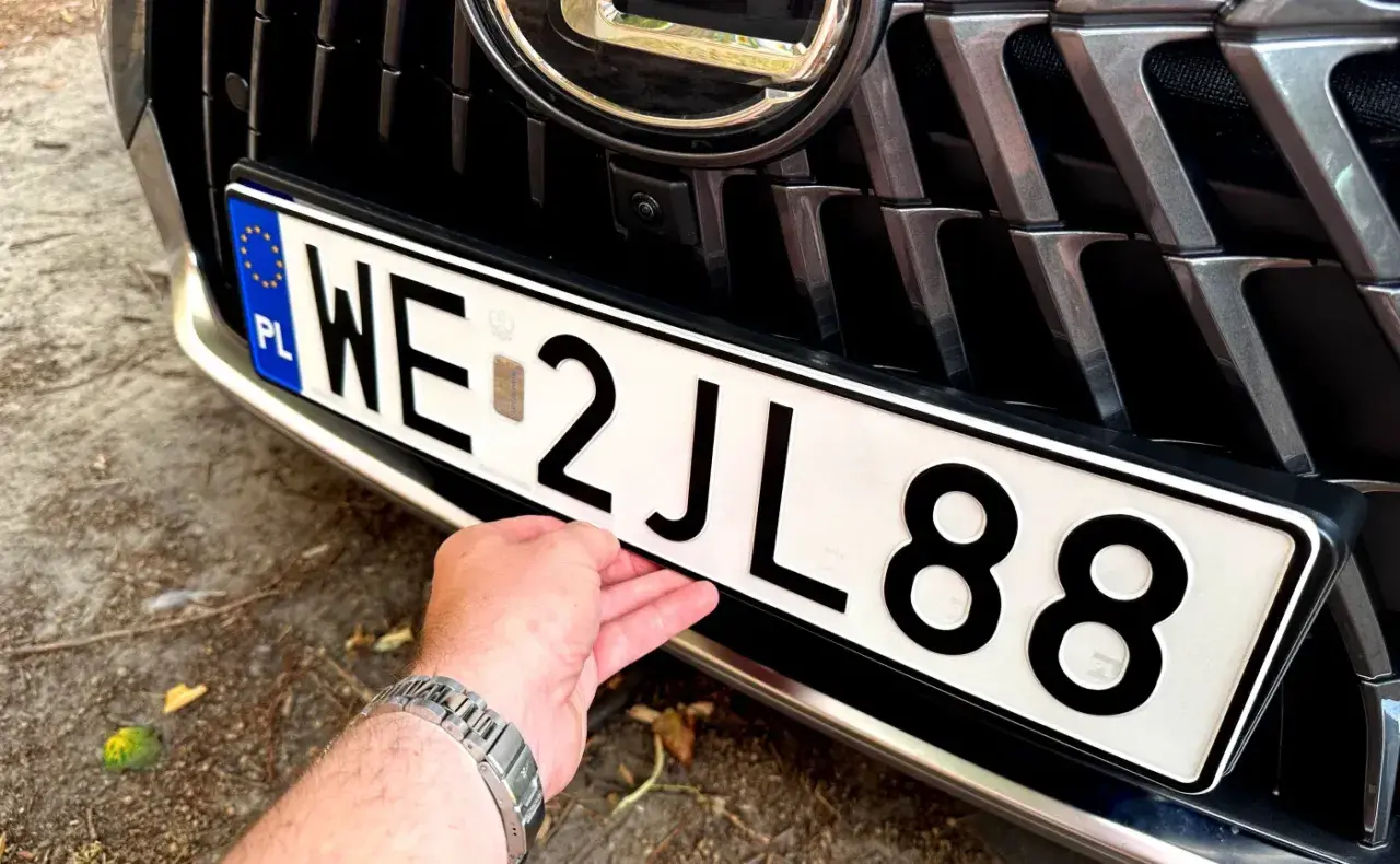 Dłoń poprawia tablicę rejestracyjną z numerem WE 2JL88.