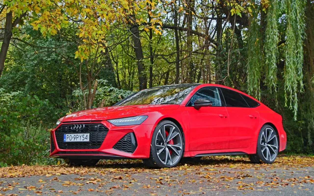 Czerwone Audi RS7 Sportback zaparkowane na tle jesiennego lasu, z liśćmi na drodze.