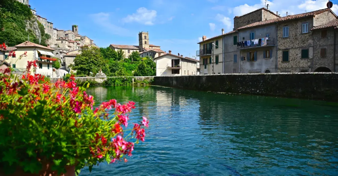 Fiori rossi in primo piano, con il borgo di Santa Fiora, Grosseto, che si specchia nell'acqua limpida.