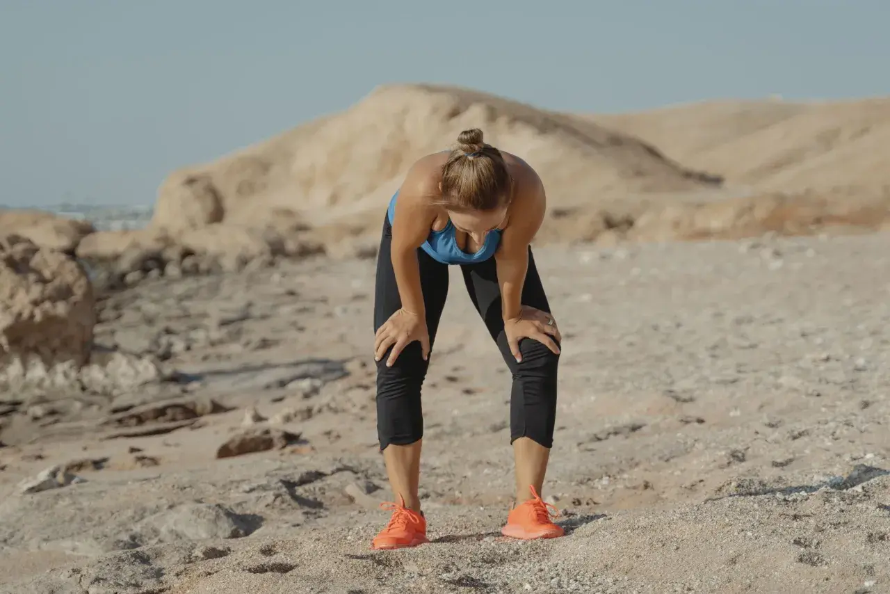 Une femme s'arrête, penchée en avant, les mains sur les genoux, visiblement fatiguée par son sport sous la canicule. Le paysage désertique accentue la chaleur.