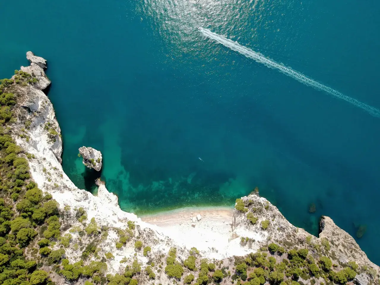 Vista aerea della cala della pipa, con scogliere bianche, vegetazione verde e mare turchese. Una barca lascia una scia sull'acqua.