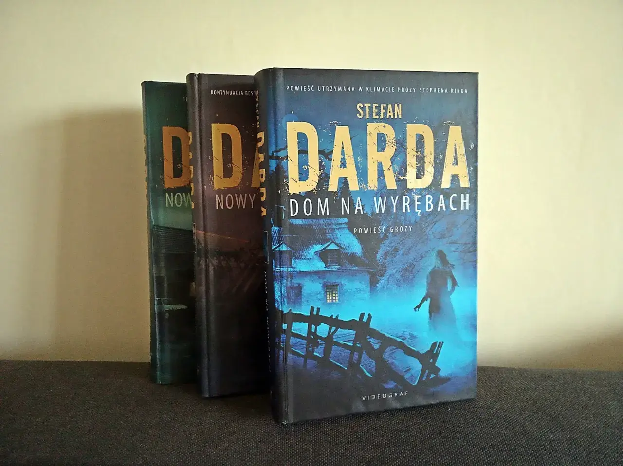 Trzy książki Stefana Dardy, w tym "Dom na wyrębach", stoją obok siebie. Okładka "Domu na wyrębach" przedstawia mroczny, niebieski krajobraz z domem i postacią.