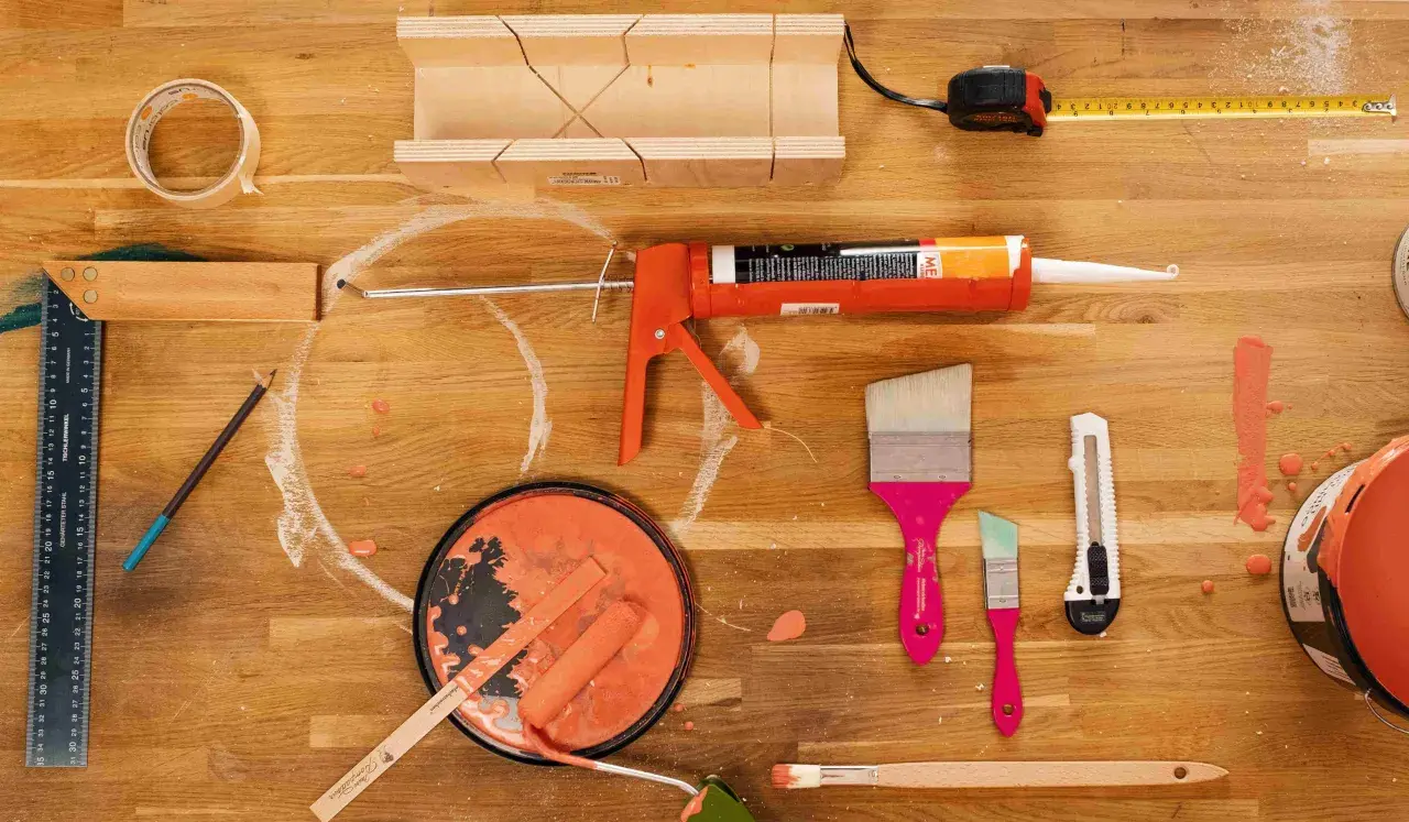 Pistolet à mastic, rouleau, pinceaux et pot de peinture orange sur un établi en bois.