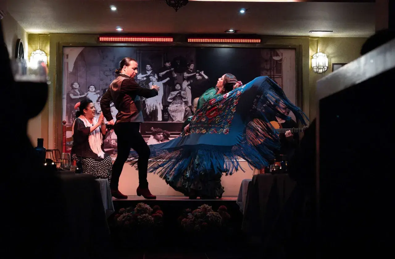 Una cantaora flamenca, con un mantón azul vibrante, baila con pasión. A su lado, un hombre la acompaña en el escenario.