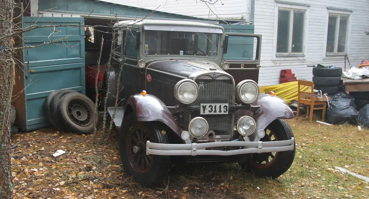 En gammal Dodge-bil står parkerad utanför ett garage. Bilen är mörkt vinröd med kromdetaljer och har en klassisk design.
