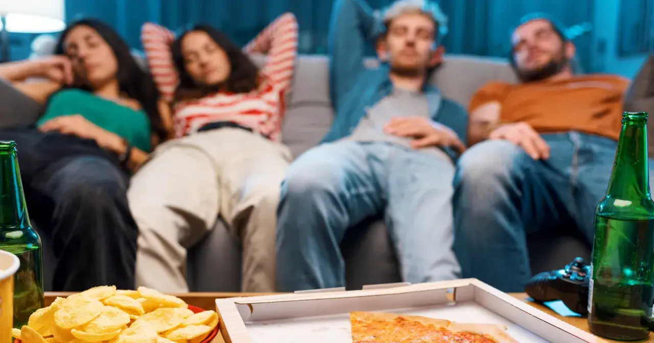 Grupa przyjaciół odpoczywa na kanapie po obfitym posiłku, czując senność. Na stole pizza, chipsy i piwo.