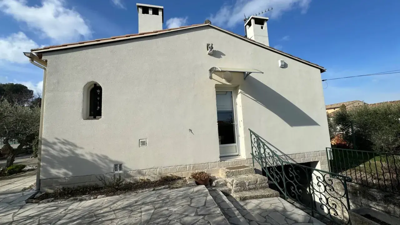 Maison Phénix 1970, blanche, avec cheminées, sous un ciel bleu. Escalier en pierre menant à la porte.