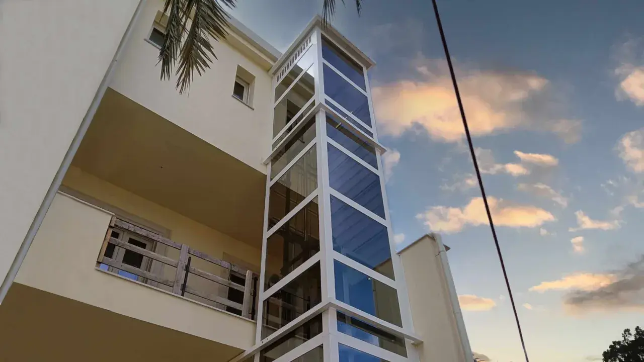 Moderno montacarichi persone esterno in vetro e metallo bianco, che sale lungo un edificio color crema, con cielo nuvoloso al tramonto.