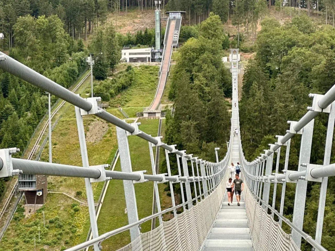 Menschen spazieren über den Willingen Skywalk, eine Hängebrücke mit Blick auf eine Skisprungschanze und bewaldete Hänge.