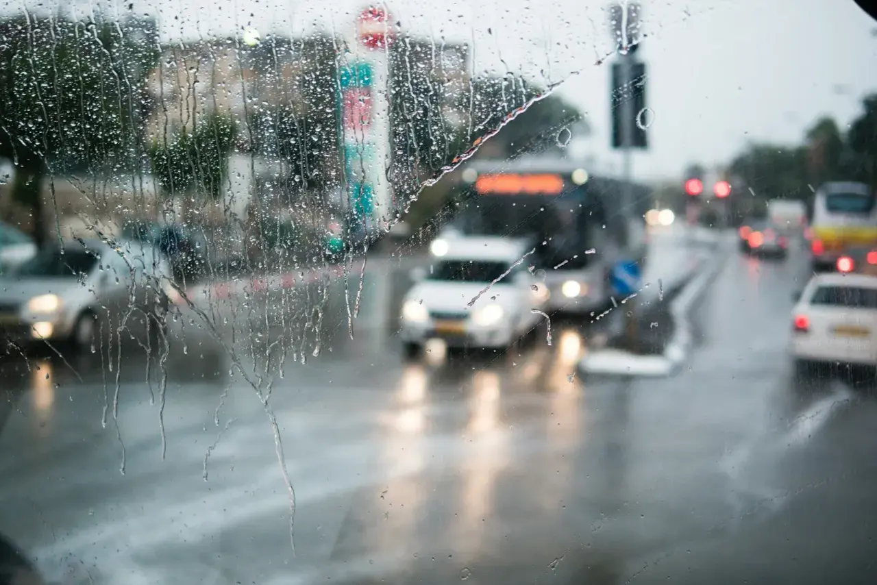 Gouttes de pluie sur un pare-brise, floutant la circulation urbaine. Les feux position seuls des voitures et des bus créent des traînées lumineuses sur la chaussée mouillée.