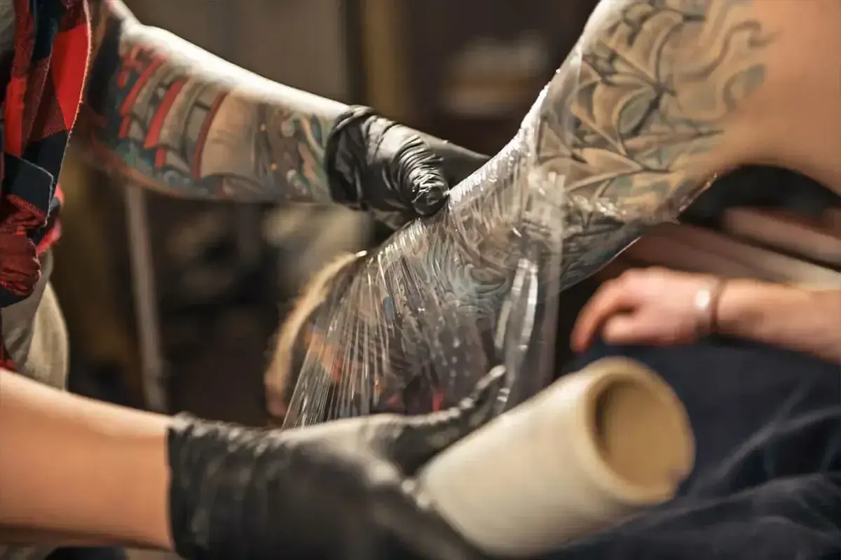 Manos enguantadas cubren un tatuaje recién hecho en el brazo con plástico. Aprende como cuidar un tatuaje recien hecho para una curación óptima.
