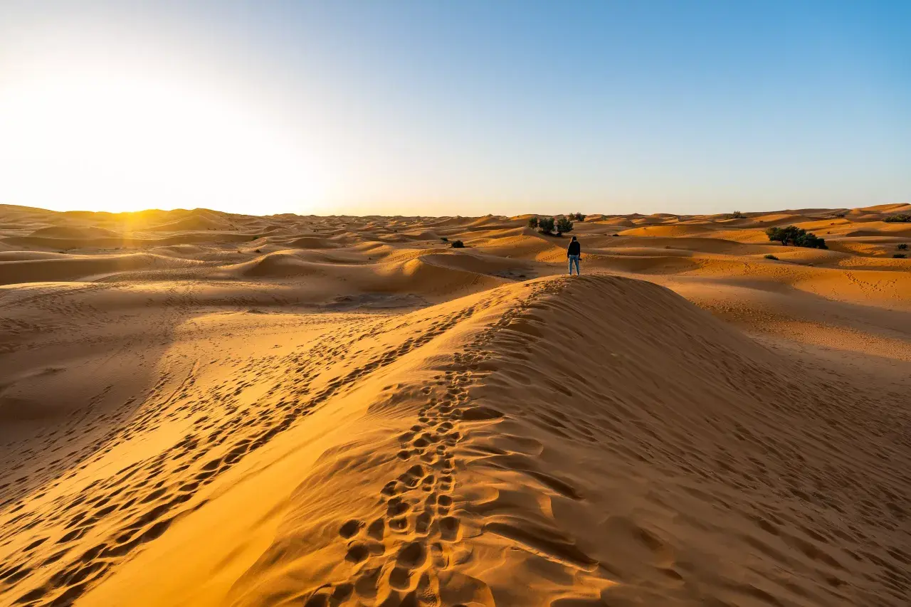 Ein einsamer Wanderer steht auf einer Düne während einer Marokko Sahara Tour. Fußspuren im Sand führen zum Horizont, wo die Sonne untergeht.