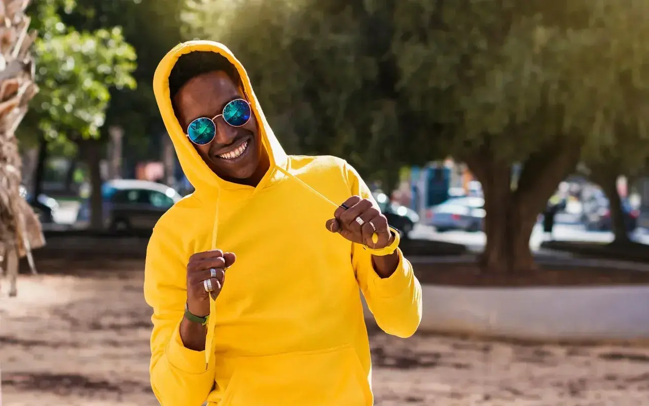 Un homme souriant porte un sweat jaune vif, ses lunettes rondes reflétant le soleil. Le grammage du sweat semble parfait pour une journée ensoleillée.
