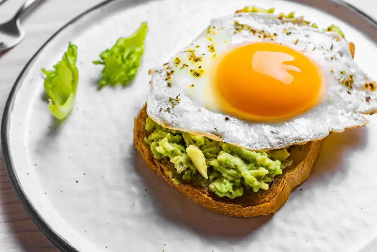 Un toast garni d'avocat écrasé et surmonté d'œufs sur le plat, assaisonné d'herbes. Deux feuilles de laitue verte décorent l'assiette.