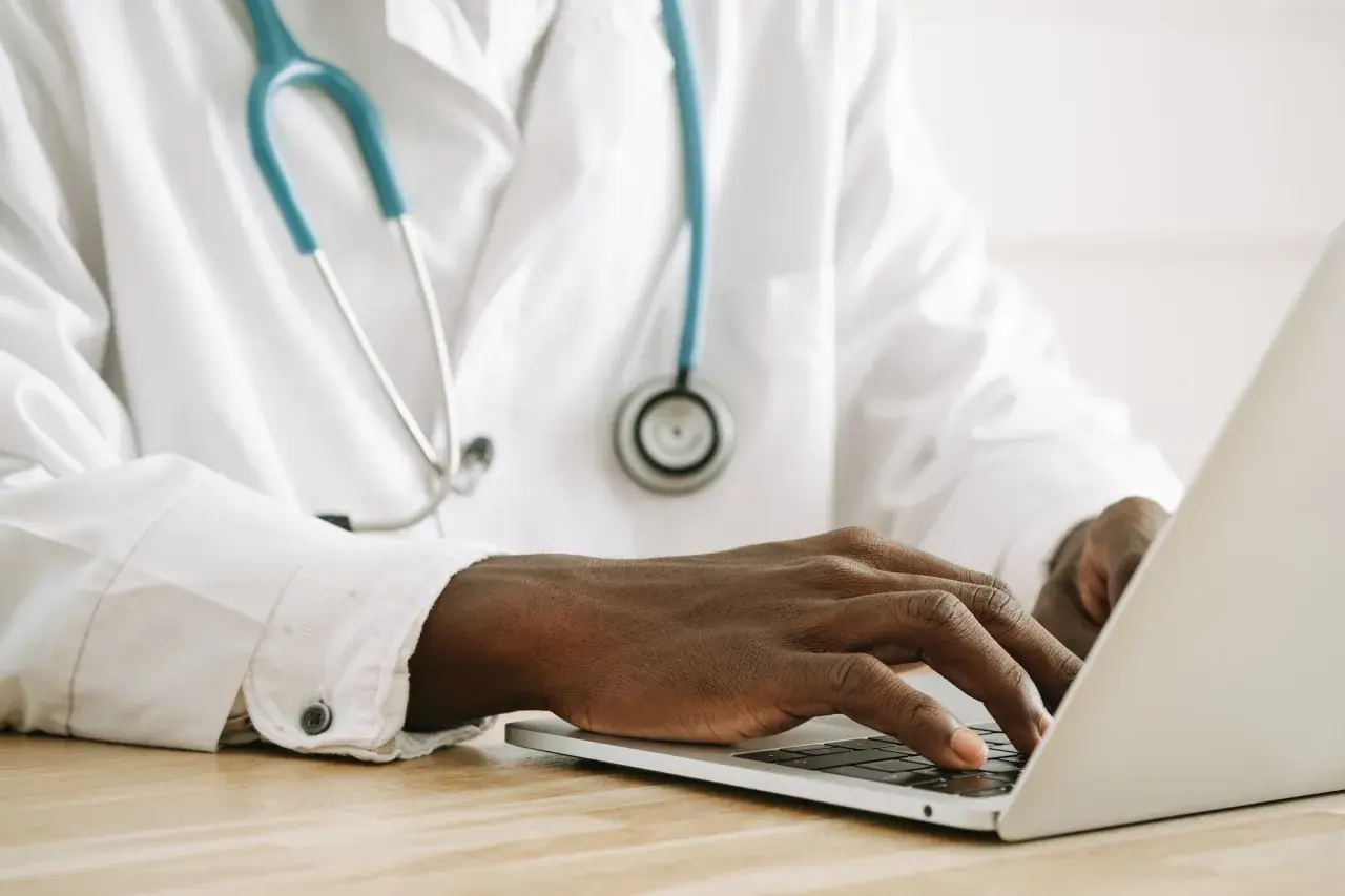 Mani di un medico che digita su un laptop, con uno stetoscopio appeso al camice bianco. Forse sta gestendo gli arretrati medici di base quando.