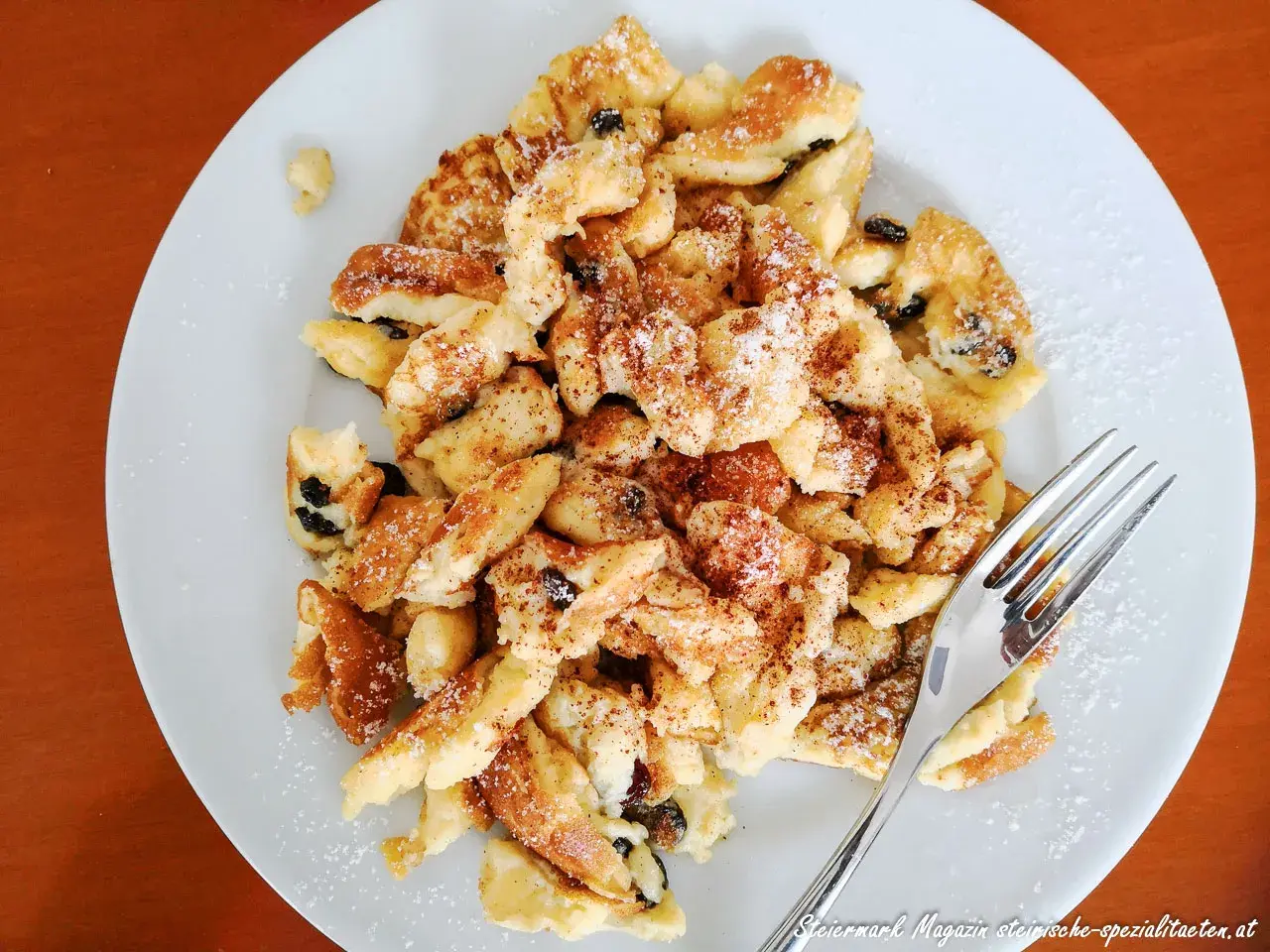 Ein Teller mit fluffigem Kaiserschmarrn mit Eischnee, bestreut mit Puderzucker und Zimt. Daneben eine Gabel.