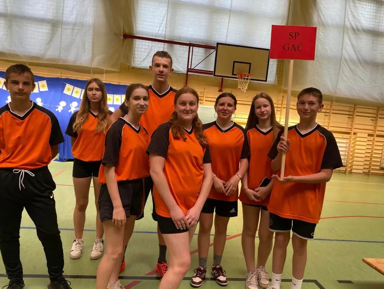 Uczniowie zespołu szkolno przedszkolnego SP GAC w pomarańczowych strojach sportowych pozują do zdjęcia w sali gimnastycznej.