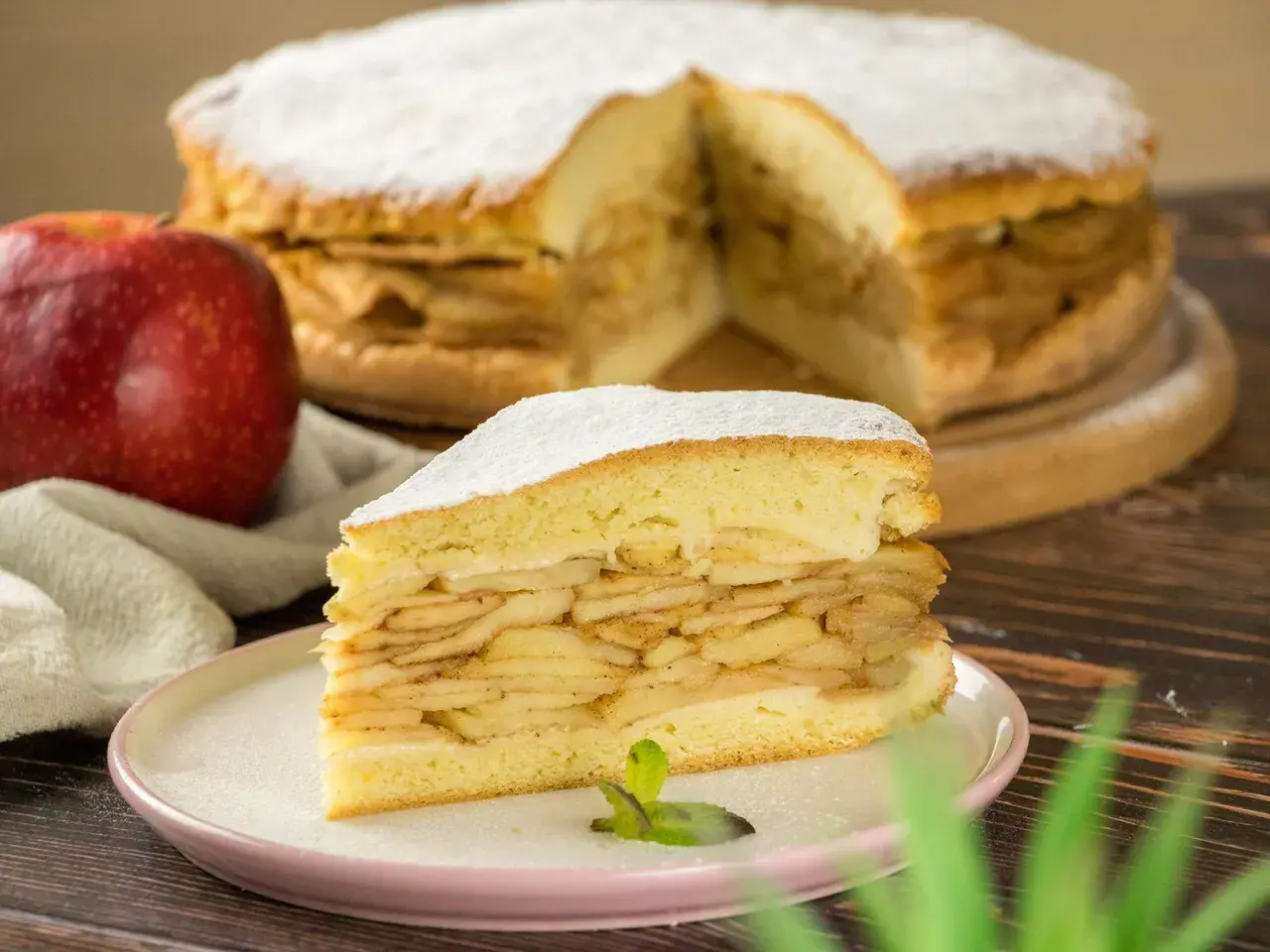 Ein saftiges Stück gedeckter Apfelkuchen mit vielen Apfelschichten, bestäubt mit Puderzucker. Daneben ein roter Apfel.