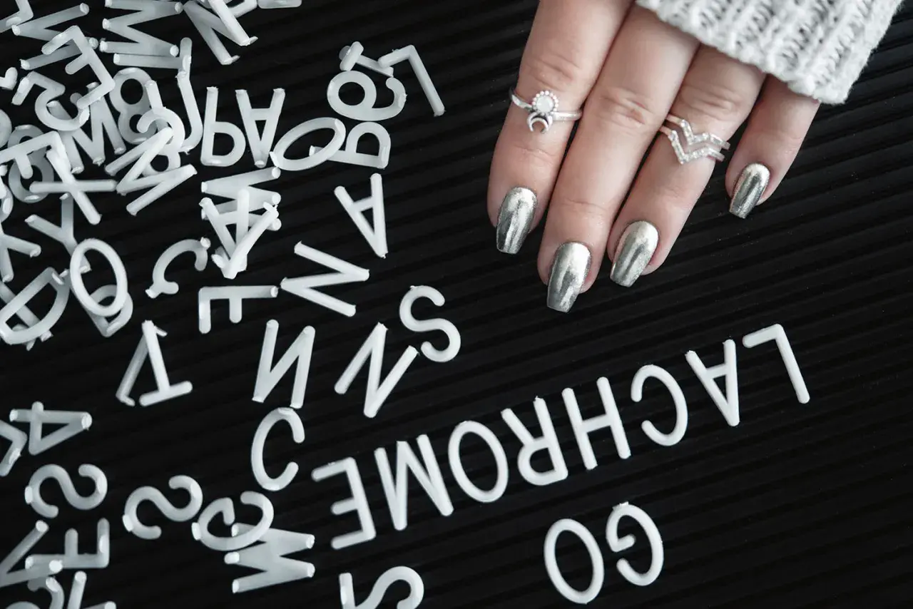 A hand with silver metallic nails and rings rests on a black surface scattered with white letters.
