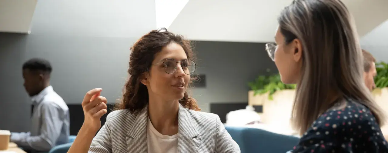 Två kvinnor i samtal, en med glasögon och lockigt hår, den andra med långt hår. En man syns i bakgrunden. Scenen utstrålar autentiskt ledarskap genom öppen kommunikation.