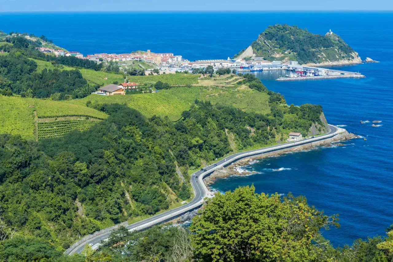 Grüne Weinberge und Wälder säumen die Küstenstraße bei San Sebastián. Das blaue Meer glitzert, während ein kleiner Hafen mit Booten in der Ferne liegt.