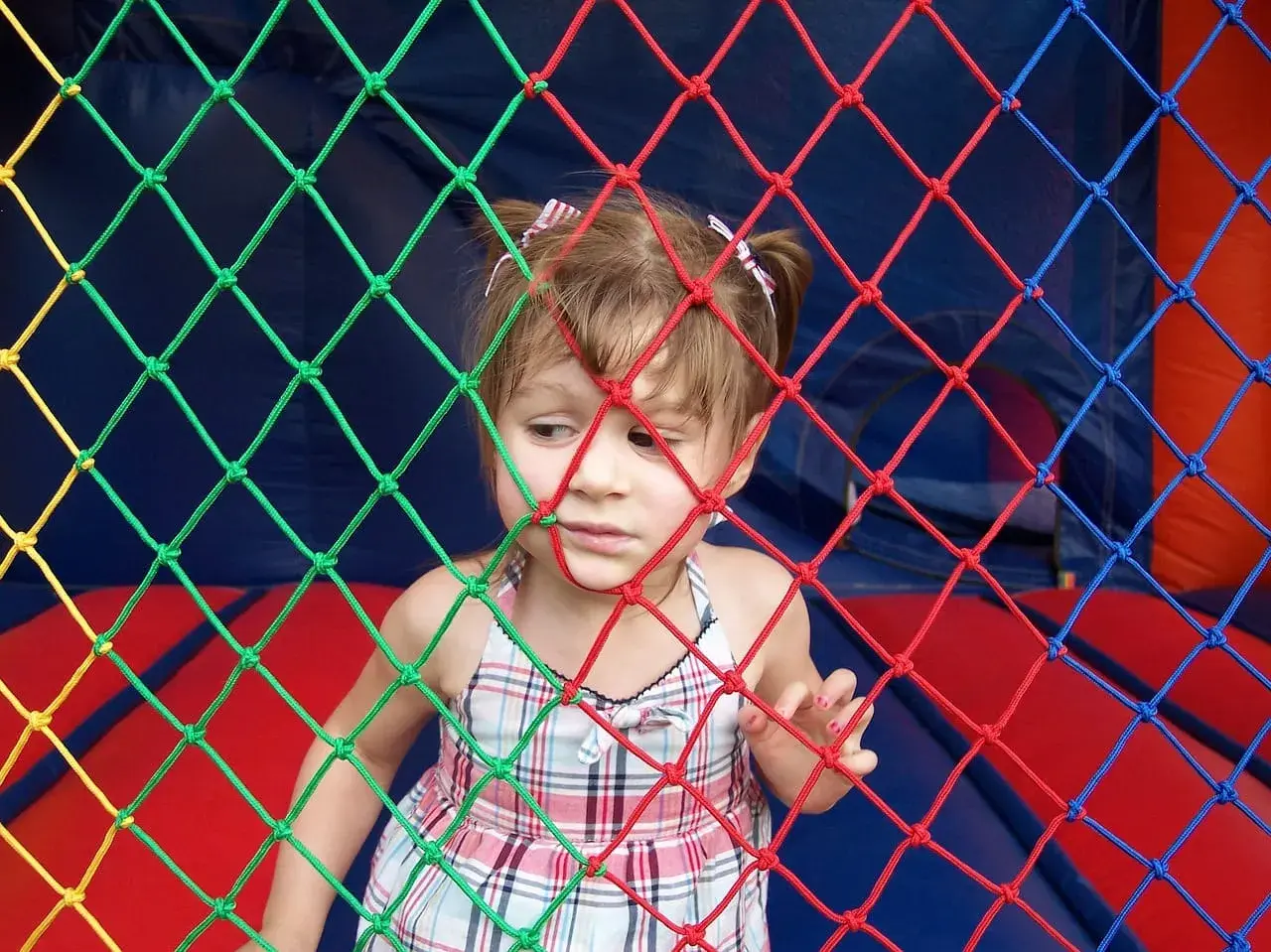 Mała dziewczynka w kratkowanej sukience patrzy przez kolorową siatkę. Może słucha piosenki dla małych dzieci.